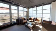 This single-floor space presents a stylish and contemporary lounge environment defined by its expansive floor-to-ceiling windows that wrap around the corner of the room, flooding the interior with abundant natural light and offering open views of the surrounding cityscape. The industrial-chic aesthetic is reinforced by an exposed concrete ceiling fitted with adjustable track lighting, providing both functional illumination and a modern design accent. The seating arrangement centers around a glass-top coffee table, surrounded by a curated mix of dark-framed armchairs with patterned cushions and wicker-style lounge chairs, creating a comfortable and inviting gathering area. The neutral gray flooring complements the overall color palette of warm tones and dark furnishings, contributing to a cohesive and professional ambiance. A structural concrete column near the window wall adds an architectural element that enhances the industrial character of the space. Frosted lower window panels provide a degree of privacy while still allowing light to permeate throughout. A potted plant near the window wall introduces a touch of greenery, softening the otherwise sleek and urban interior. This space is well-suited for collaborative meetings, casual gatherings, or relaxed work sessions.