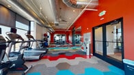 This fitness center makes a strong visual statement through its bold design choices and comprehensive equipment offerings. The most immediately striking element is the vivid red accent wall, which anchors the space and sets an energetic tone throughout. Complementing this is the eye-catching hexagonal tile flooring, featuring an interlocking pattern of red, teal, yellow, gray, and blue tiles that adds dynamic visual interest underfoot.

The ceiling draws the eye upward, where exposed silver ductwork runs the length of the space alongside recessed lighting and ceiling fans, lending an industrial aesthetic that balances the colorful design elements below. Large floor-to-ceiling windows line one wall, flooding the interior with abundant natural light and offering open views of the surrounding neighborhood.

The layout is thoughtfully organized into distinct functional zones. A dedicated cardio area houses treadmills, ellipticals, and stationary bikes positioned to take advantage of the expansive window views. A strength training zone features multi-use cable machines and a weight rack, while a free-weight area offers dumbbells, medicine balls, stability balls, and exercise mats for versatile workouts. A glass-paneled partition wall partially divides sections of the space, adding architectural structure while maintaining an open, airy feel throughout.