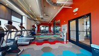 This fitness center makes a strong visual statement through its bold design choices and comprehensive equipment offerings. The most immediately striking element is the vivid red accent wall, which anchors the space and sets an energetic tone throughout. Complementing this is the eye-catching hexagonal tile flooring, featuring an interlocking pattern of red, teal, yellow, gray, and blue tiles that adds dynamic visual interest underfoot.

The ceiling draws the eye upward, where exposed silver ductwork runs the length of the space alongside recessed lighting and ceiling fans, lending an industrial aesthetic that balances the colorful design elements below. Large floor-to-ceiling windows line one wall, flooding the interior with abundant natural light and offering open views of the surrounding neighborhood.

The layout is thoughtfully organized into distinct functional zones. A dedicated cardio area houses treadmills, ellipticals, and stationary bikes positioned to take advantage of the expansive window views. A strength training zone features multi-use cable machines and a weight rack, while a free-weight area offers dumbbells, medicine balls, stability balls, and exercise mats for versatile workouts. A glass-paneled partition wall partially divides sections of the space, adding architectural structure while maintaining an open, airy feel throughout.