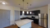 This single-floor space is centered around a well-appointed kitchen that blends modern aesthetics with functional design. The kitchen serves as the heart of the space, featuring rich dark cabinetry that extends to the upper walls, providing ample storage while creating a bold, sophisticated contrast against the crisp white subway tile backsplash. A suite of stainless steel appliances — including a range, microwave, dishwasher, and refrigerator — ensures a fully equipped cooking environment. A central island with a light-toned granite countertop offers generous workspace and serves as a natural focal point, ideal for casual dining or meal preparation. Three pendant lights hang above the island, casting warm, focused light and adding a contemporary decorative touch. A track lighting fixture provides additional ambient illumination. The warm hardwood flooring grounds the space and complements the overall color palette. Light-toned walls keep the atmosphere bright and open. The layout is efficient and thoughtfully organized, with clear sightlines throughout the kitchen that contribute to a sense of openness. Overall, the space delivers a clean, modern ambiance with quality finishes throughout.