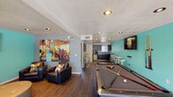 This single-floor recreational space is designed for socializing and entertainment, combining a lively game room with a comfortable lounge area and a functional kitchenette. The open layout is anchored by a full-size pool table positioned at the center, accompanied by a wall-mounted cue rack and modern bar-height tables with contemporary pedestal stools along the bold teal accent wall. Two wall-mounted flat-screen TVs ensure entertainment from multiple vantage points throughout the room. The lounge area features dark leather seating — including a sofa, armchairs, and a tufted ottoman — arranged around one of the screens, creating an inviting space to relax. A large, colorful piece of artwork adds an energetic, urban aesthetic to the lounge corner. The kitchenette is outfitted with dark cabinetry, a mosaic tile backsplash, stainless steel sink, compact refrigerator, and a coffee station, making it well-suited for hosting. Rich hardwood-style flooring runs throughout, complementing the textured ceiling with recessed lighting. Large windows and a glass door provide abundant natural light and direct views and access to an outdoor area, seamlessly connecting the interior to the exterior environment.