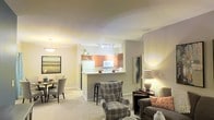 This single-floor residence offers a thoughtfully designed open-concept layout that seamlessly connects the main living areas, creating a warm and inviting atmosphere throughout. The living area is carpeted and comfortably sized, providing an ideal setting for relaxation and everyday living, with warm ambient lighting that adds to the cozy feel of the space. A plaid armchair and upholstered seating anchor the room, complemented by tasteful wall art and accent lighting.

Adjacent to the living area, a dedicated dining space features an elegant pendant light overhead, with seating for several guests — perfect for both casual meals and entertaining. The two areas flow naturally into one another, enhancing the open, connected feel of the floor plan.

The kitchen is positioned at the heart of the home, defined by a peninsula with bar stool seating that serves as both a functional workspace and a social hub. Warm wood-toned cabinetry, a white refrigerator, and a built-in microwave outfit the kitchen with everyday essentials. Track lighting illuminates the workspace efficiently. A wine rack above the upper cabinets adds a charming, practical touch. The smooth, neutral-toned walls and consistent flooring throughout the space contribute to a cohesive and harmonious interior aesthetic.