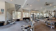 Located at 6850 Peachtree Dunwoody Rd in Sandy Springs, GA, this fitness center offers a comprehensive workout facility spread across a single floor with access to an upper level via a striking spiral staircase. The main gym area is generously sized and flooded with natural light through multiple large windows that frame views of surrounding greenery, creating a bright and energizing atmosphere. The space is carpeted throughout the workout zones, providing comfort underfoot during training sessions. Full-length mirror walls line one side of the strength training area, visually expanding the space while allowing users to monitor their form. The strength training section is well-appointed with a multi-station cable machine, free weights including a complete dumbbell rack, weight benches, and a squat/power rack. A dedicated cardio zone houses treadmills and elliptical trainers, each positioned in front of wall-mounted flat-screen TVs and large windows overlooking the outdoors. Recessed lighting and ceiling fans contribute to a comfortable and well-lit environment. The upper level, accessible via the spiral staircase, features bamboo hardwood flooring, high ceilings, a ceiling fan, and French doors that open to an outdoor area, offering a versatile space ideal for stretching, yoga, or light exercise.