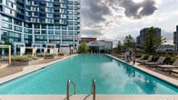 Located in McLean, Virginia, this outdoor amenity space offers a resort-inspired rooftop pool experience framed by the striking modern high-rise tower above. The centerpiece is a generously proportioned swimming pool with crystal-clear turquoise water, clean tile surrounds, and multiple entry points including stainless steel handrails. The pool deck is thoughtfully designed with ample space for lounging, featuring rows of contemporary reclining chairs and shaded private cabanas draped in white curtains for more secluded relaxation. Lush landscaping integrates seamlessly throughout the space, with neatly trimmed shrubs, ornamental trees, and planted garden beds adding greenery and texture. A dedicated sun terrace extends beyond the pool area, offering additional lounge seating with open-sky exposure and panoramic views of the McLean and Tysons skyline, including neighboring office towers and urban infrastructure. The elevated vantage point provides a dynamic backdrop that transitions from daytime brightness to dramatic cloudy skies. Utility structures and service areas are discreetly incorporated into the layout. Overall, the space combines modern architectural design with refined outdoor living amenities, creating an inviting and upscale environment ideal for relaxation and recreation.