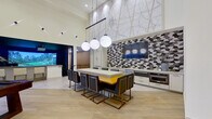This single-floor amenity space at The Huntley is thoughtfully designed to deliver a premium social and recreational experience. The layout encompasses several distinct zones, each with its own character while maintaining a cohesive contemporary aesthetic throughout.

The main lounge area commands attention with dramatically soaring ceilings and an abundance of natural light streaming through expansive floor-to-ceiling windows. Clean white walls are accented by geometric wall-mounted lighting fixtures, and light wood-style flooring runs throughout, lending warmth to the open atmosphere. Comfortable modern sofas with colorful throw pillows provide inviting seating, while views to an outdoor area enhance the sense of openness.

A dedicated billiards zone features a handsome dark-felt pool table positioned beneath high clerestory windows that frame outdoor views of the surrounding building courtyard.

Adjacent to the billiards area, a fully equipped kitchenette serves as a social hub, anchored by a large island with bold gold-and-black bar seating. Branded globe pendant lights hang overhead, and a striking geometric-patterned tile backsplash frames a wall-mounted television. A large vintage-style map mural adorns the upper wall, adding a distinctive local touch.

The standout feature is a dedicated golf simulator room with dark acoustic wall panels, professional-grade turf mats, and a large projection screen delivering an immersive on-course experience.