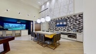 This single-floor amenity space at The Huntley is thoughtfully designed to deliver a premium social and recreational experience. The layout encompasses several distinct zones, each with its own character while maintaining a cohesive contemporary aesthetic throughout.

The main lounge area commands attention with dramatically soaring ceilings and an abundance of natural light streaming through expansive floor-to-ceiling windows. Clean white walls are accented by geometric wall-mounted lighting fixtures, and light wood-style flooring runs throughout, lending warmth to the open atmosphere. Comfortable modern sofas with colorful throw pillows provide inviting seating, while views to an outdoor area enhance the sense of openness.

A dedicated billiards zone features a handsome dark-felt pool table positioned beneath high clerestory windows that frame outdoor views of the surrounding building courtyard.

Adjacent to the billiards area, a fully equipped kitchenette serves as a social hub, anchored by a large island with bold gold-and-black bar seating. Branded globe pendant lights hang overhead, and a striking geometric-patterned tile backsplash frames a wall-mounted television. A large vintage-style map mural adorns the upper wall, adding a distinctive local touch.

The standout feature is a dedicated golf simulator room with dark acoustic wall panels, professional-grade turf mats, and a large projection screen delivering an immersive on-course experience.