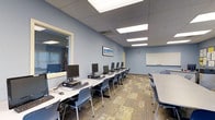 The Computer Learning Center is a single-floor educational facility thoughtfully designed to support both individual and group learning experiences. The main classroom features a row of desktop computer workstations arranged along a long white table, each station equipped with a monitor, keyboard, and mouse — providing ample space for hands-on digital instruction. Additional seating is available at a separate table to the right, accommodating collaborative work or observation. A large whiteboard mounted on the wall serves as a central focal point for instruction, making the room well-suited for guided lessons and presentations. The space is finished with blue-toned walls that create a calm, focused atmosphere, complemented by a patterned carpet in warm neutral tones that adds texture underfoot. Recessed ceiling panels with flush fluorescent lighting ensure the room is brightly and evenly illuminated throughout. A framed artwork piece on the wall adds a touch of character to the learning environment. An adjacent windowed area, visible through an interior window, suggests additional functional space nearby. The overall layout is practical and organized, optimized for technology-driven education in a clean, professional setting.