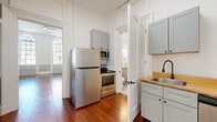 This single-floor unit showcases a compelling mix of historic architectural character and contemporary updates throughout its well-proportioned layout. The living room immediately impresses with soaring ceilings, gleaming wide-plank hardwood floors, and tall arched windows that flood the space with abundant natural light. Two-tone walls featuring gray wainscoting paired with crisp white upper walls reinforce the classic aesthetic, while ornate door casings and transom details reflect the building's historic origins.

Multiple hallways connect the rooms in an efficient, flowing arrangement. The kitchen is thoughtfully equipped with stainless steel appliances—including a refrigerator, electric range, microwave, and dishwasher—complemented by gray shaker-style cabinetry, warm butcher block countertops, and a matte black faucet over a stainless sink. Rich hardwood floors extend through the kitchen, maintaining visual continuity.

The bathroom offers a contrasting palette, with light wood-look tile flooring, a gray vanity with butcher block countertop, and clean white walls, creating a bright, airy feel. Throughout the unit, tall ceilings, generous millwork, and an abundance of natural light contribute to a spacious and distinguished ambiance.