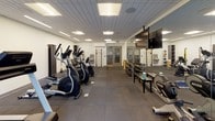 This modern fitness center is thoughtfully designed to support a comprehensive workout experience within a single open floor plan. The space is finished with durable dark rubber flooring tiles throughout, providing both safety and practicality for a wide range of exercises. Recessed panel lighting in the suspended ceiling ensures the entire area is evenly and brightly illuminated.

A standout architectural feature is the full-width glass garage-style door along one wall, which floods the cardio zone with natural light and offers views to an outdoor area, creating an open, airy atmosphere. Full-length mirrors are positioned strategically along multiple walls, visually expanding the space and supporting proper form during workouts.

The cardio section is well-stocked with a row of Spirit-brand treadmills, elliptical trainers, and stationary bikes. The free weights area houses a tiered dumbbell rack with a comprehensive weight selection, accompanied by benches for strength training. Additional equipment includes a cable machine, TRX suspension straps, a wall-mounted pull-up and ladder system, and a medicine ball rack. A wall-mounted flat-screen television is positioned for visibility across the cardio zone. The space is completed with water stations and sanitation supplies, reflecting a focus on user convenience and hygiene.