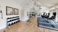 Located at 107 Felix St in Santa Cruz, California, this single-floor fitness center is designed to provide a bright, well-equipped workout environment. The space is anchored by striking vaulted ceilings with exposed white-painted beams, giving the interior a sense of generous vertical openness. Warm wood-look flooring runs throughout, complementing the clean white walls and creating an inviting, airy atmosphere.

The cardio zone features a row of modern treadmills and elliptical trainers, all equipped with digital displays, positioned along a wall of large glass doors and transom windows that frame lush outdoor greenery and flood the space with natural light. A ceiling fan and multiple pendant-style barn lights provide additional ventilation and warm illumination throughout.

On the strength training side, the facility offers a full functional training rig complete with TRX suspension straps, kettlebells, and resistance attachments, alongside a well-stocked dumbbell rack with a flat utility bench. Floor-to-ceiling mirrors visually expand the space and allow for proper form checks during workouts.

Additional amenities include a water cooler station, a fire extinguisher, and framed Santa Cruz-themed artwork that adds local character to the space. An exterior door provides convenient outdoor access, enhancing the overall sense of connectivity with the surrounding environment.