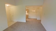 This single-floor residence presents an open and functional layout with a seamless flow between the main living area, dining space, and kitchen. The living room features neutral beige carpet and light-toned walls, creating a bright and understated atmosphere. The entry area transitions from tile flooring into the carpeted living space, delineating zones naturally. A wide pass-through opening connects the living room to the dining area, which is highlighted by a classic multi-bulb chandelier that adds a modest decorative touch. The dining area shares its open space with easy access to the kitchen, which features light wood cabinetry, a visible sink, and what appears to be a full-size refrigerator. The kitchen offers a clean, practical design with ample cabinet storage. Throughout the space, white walls and neutral carpeting maintain a consistent, cohesive aesthetic. The high ceiling line in the living area contributes to an airy sense of volume. Overall, the layout is straightforward and well-connected, making efficient use of the available space while maintaining a clean, move-in-ready appearance.