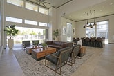 Located at 1108 Ponderosa Dr in Petaluma, California, this contemporary single-floor community space offers an impressive blend of modern design and welcoming functionality. The main lounge commands immediate attention with its soaring double-height ceilings and a dramatic floor-to-ceiling dark metal fireplace surround that serves as a striking architectural centerpiece. Large clerestory windows flood the space with abundant natural light while offering views of the surrounding community. The living area is furnished with tufted leather chairs and rich wood-accented seating arrangements set atop neutral stone tile flooring, creating a refined yet comfortable atmosphere. A separate lounge nook features warm cognac leather wingback chairs, gold-toned accent tables, and a wall-mounted TV, ideal for casual gatherings. Organic artwork, including a sculptural wood-grain wall piece, adds a natural, artistic touch throughout. The communal kitchen and dining area is appointed with an expansive stainless steel island, dark wood cabinetry, and a dramatic cluster of black-and-gold pendant lights hanging above leather-upholstered bar seating. Large windows brighten the kitchen, maintaining the space's open, airy character. Together, these areas form a cohesive, elegantly appointed community hub designed for both social interaction and relaxed everyday use.