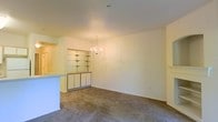 This single-floor space offers a clean and functional layout designed for comfortable everyday living. The main living and dining area is open and airy, finished with neutral-toned carpet that extends throughout the space, lending a warm and cohesive feel underfoot. A classic chandelier serves as a central decorative focal point, providing both ambient lighting and a touch of elegance to the dining area.

A built-in shelving unit with a lower cabinet base is integrated into one wall, offering ample display and storage opportunities without sacrificing floor space. This feature adds a practical yet architectural element to the room's design. Adjacent to this area, a kitchen bar counter provides a natural transition between the cooking and living spaces, making the layout well-suited for casual dining or entertaining.

The walls are finished in a soft, light tone that reflects available light and contributes to the overall bright and open ambiance of the space. The ceiling is smooth and uncluttered, reinforcing the clean aesthetic. Overall, the space presents a straightforward and adaptable interior with thoughtful built-in details that enhance both its functionality and visual character.