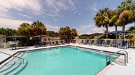 This outdoor pool facility in Pensacola, Florida offers a well-designed recreational space ideal for relaxation and leisure. At the heart of the space is a generously sized swimming pool with crystal-clear turquoise water, surrounded by a broad, light-colored concrete deck that provides ample room for lounging. Multiple reclining chaise lounges are arranged along the poolside, offering convenient spots to soak up the Florida sun.

The deck area is further enhanced by several dining-height tables paired with chairs, each shaded by large navy blue cantilever umbrellas, providing comfortable areas for outdoor seating and socializing. The perimeter of the pool deck is enclosed by a white picket-style fence, lending a clean, classic aesthetic to the space.

Adjacent to the pool deck is a single-story support structure featuring a charming combination of white lap siding and stacked stone accents. Classic column details and wall-mounted lantern lighting contribute to the building's coastal character. French-style doors with louvered blinds provide access to the interior amenity space.

The entire area is beautifully framed by mature tropical landscaping, including tall palm trees and lush pines, creating a private, resort-like atmosphere. Open skies above allow for abundant natural light, making this pool area a bright and inviting outdoor destination.