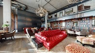 This striking single-floor space masterfully blends industrial architecture with refined, eclectic interior design. Soaring ceilings with exposed concrete and visible ductwork set a dramatic tone, while riveted steel wall panels and structural columns reinforce the raw, industrial character of the space. Overhead, elegant crystal chandeliers create a compelling contrast, casting warm light across wide-plank wood flooring and layered area rugs below.

The main lounge area is anchored by a commanding tufted red leather Chesterfield sofa, surrounded by an eclectic mix of wingback chairs, leather seating, and vintage-style furnishings. A linear fireplace provides a warm focal point, while floor-to-ceiling built-in bookshelves line the walls, filled with books and decorative objects. A mezzanine level overlooks the main floor, adding vertical depth and architectural interest.

A separate entry lounge features expansive floor-to-ceiling windows that flood the space with natural light and offer views of the surrounding urban environment. Dramatic large-scale artwork and textured faux-finish walls adorn the corridors, contributing to the gallery-like atmosphere.

A bar area with brick surround, pendant lighting, and a mounted display screen serves as another key gathering point. Throughout, the space maintains a sophisticated, club-like ambiance defined by bold design choices and rich material contrasts.