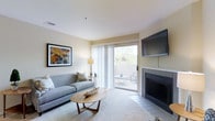 This single-floor space offers a thoughtfully designed living environment with a calm, contemporary aesthetic. The living room serves as the heart of the space, featuring soft neutral wall tones and plush carpeting that create a warm, welcoming atmosphere. A tile-surround fireplace anchors one side of the room, providing both a functional and visual focal point, complemented by a wall-mounted flat-screen television above. Large sliding glass doors flood the interior with natural light and open directly onto a private outdoor deck, where glimpses of surrounding greenery offer a serene connection to the outdoors. The deck itself provides an inviting extension of the living area, ideal for relaxing or casual outdoor enjoyment. Decorative touches, including colorful wall art and carefully chosen furnishings such as a contemporary sofa and a distinctive oval wood coffee table, add personality and style to the space. Recessed lighting in the ceiling contributes to an even, clean illumination throughout the room. Overall, the space balances comfort and modern design sensibility, making it a functional and visually appealing environment suited to everyday living.