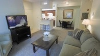 This single-floor residence features an open-concept design that seamlessly integrates the living, dining, and kitchen areas into one cohesive space. The living room is anchored by a plush sofa and a dark wood coffee table topped with a floral centerpiece, with a flat-screen TV positioned on a matching media console along the wall. Neutral-toned carpeting runs throughout the main living and dining areas, contributing to a warm and inviting ambiance.

The kitchen is set slightly apart, defined by a raised breakfast bar with two bar stools, offering both a casual dining option and a natural visual divider between the cooking and living spaces. Medium-toned wood cabinetry lines the upper kitchen walls, complemented by light countertops and a white refrigerator. Tile flooring in the kitchen contrasts neatly with the carpeted areas beyond.

Adjacent to the living room, a compact dining area accommodates a dark round table with matching chairs, lit by overhead lighting and accented by a framed wall piece. The overall color palette is soft and neutral, with beige walls and consistent finishes creating a cohesive, functional living environment throughout.