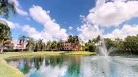 This single-story residence is nestled within a beautifully landscaped tropical community that immediately impresses with its picturesque outdoor surroundings. The exterior setting is defined by a expansive, tranquil lake positioned at the forefront of the property, complete with an elegant decorative fountain that adds movement and visual interest to the waterscape. Towering royal palms and a variety of mature tropical trees frame the scene, creating a verdant, resort-like atmosphere that is characteristic of South Florida living.

The building itself features a warm terracotta and earth-toned facade, consistent with Mediterranean-influenced architecture commonly found in the region. Covered balconies and generous fenestration are visible on the structure, suggesting an emphasis on capturing natural light and outdoor views throughout the interior. Well-manicured lawns and lush greenery surround the property, enhancing its tropical character.

The community's overall ambiance is calm and scenic, with the lake and fountain serving as a central visual anchor. The combination of tropical landscaping, waterfront positioning, and warm architectural tones creates an inviting and relaxed environment that reflects the natural beauty of its South Florida setting.