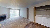 Located at 11328 SE Kent Kangley Rd in Kent, Washington, this ground-floor unit offers a practical and comfortable single-level layout. The living room serves as the heart of the home, featuring plush carpeting, a tile-surround wood-burning fireplace, and a large sliding glass door fitted with vertical blinds that invites natural light and provides access to the outdoors. Baseboard heating is present throughout, ensuring consistent warmth. The galley-style kitchen is thoughtfully appointed with warm wood-toned cabinetry, light countertops, a double sink, and white appliances including a range, refrigerator, and dishwasher, all set atop durable wood-look flooring. A dedicated in-unit laundry closet with a washer is conveniently accessible from the main living area. The bedrooms are carpeted and well-sized, each offering closet storage — one with sliding wood-panel doors — and windows that allow natural light and exterior views. The bathroom features wood-toned cabinetry, a single-basin vanity with generous counter space, a large wall mirror with overhead lighting, and wood-look flooring. Textured ceilings are consistent throughout the unit, contributing to its classic residential character.
