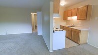 This single-floor residence offers a practical and well-organized layout designed for comfortable everyday living. The kitchen is equipped with warm honey-toned wood cabinetry providing ample storage, a white electric coil range, and a double-basin sink set beneath upper cabinets. Light-toned countertops and a light flooring surface give the kitchen a clean, bright appearance. A partial wall divider neatly separates the kitchen from the adjoining living area, which features wall-to-wall carpeting in a neutral tone that contributes to a cozy and inviting atmosphere. The open sightline between the kitchen and living space creates a connected feel throughout the main area. A hallway visible from the main living space leads further into the unit, offering access to additional rooms, including what appears to be a bedroom with a closet and a bathroom. Overhead lighting is present throughout, ensuring adequate illumination across all areas. The overall color palette is light and neutral, with beige and cream tones dominating the walls, floors, and fixtures, lending the space a clean and move-in-ready character.