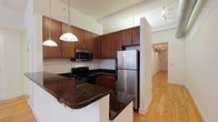 The Calder is a thoughtfully designed two-bedroom, two-bathroom residence that offers generous single-floor living across a substantial footprint. The kitchen serves as a central highlight of the home, featuring rich warm-toned cabinetry that extends to the ceiling for ample storage. Dark granite countertops provide a striking contrast against the cabinetry, while a breakfast bar peninsula offers additional prep space and casual seating. Stainless steel appliances, including a full-size refrigerator and built-in microwave, complement the sleek aesthetic, and pendant lighting adds a polished finishing touch overhead. Hardwood flooring flows from the kitchen into the hallway and living areas, contributing warmth and continuity throughout the space. The hallway leading to the bedrooms is well-lit and spacious, suggesting comfortable room separation and privacy. Track lighting in the connecting corridor enhances the sense of openness. With two full bathrooms, the layout accommodates both residents and guests with ease. The overall ambiance balances modern design elements with functional living spaces, making The Calder a well-appointed and practical home.