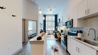 This single-story unit showcases a striking open-concept layout defined by dramatically tall ceilings that amplify the sense of space throughout. The galley-style kitchen runs along one wall and is outfitted with white shaker cabinetry, granite countertops, a subway tile backsplash, and a full suite of stainless steel appliances including a refrigerator, electric range, microwave, and dishwasher. A peninsula counter extends into the living area, providing additional workspace and casual seating.

The living area sits at the far end of the open floor plan, anchored by oversized windows dressed with teal drapes that draw in generous natural light and offer outdoor views. A ceiling fan overhead adds functionality to the airy volume. Wood-style plank flooring flows consistently throughout the main living spaces, grounding the bright white walls and high ceilings.

The bedroom features tall windows that continue the theme of abundant natural light, with high ceilings creating a spacious feel. The full bathroom includes a granite vanity, white cabinetry, wood-style flooring, and a tiled tub-shower combination. A dedicated laundry area houses a stacked washer and dryer unit alongside built-in shelving for storage, completing this well-appointed space.