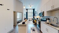 This single-story unit showcases a striking open-concept layout defined by dramatically tall ceilings that amplify the sense of space throughout. The galley-style kitchen runs along one wall and is outfitted with white shaker cabinetry, granite countertops, a subway tile backsplash, and a full suite of stainless steel appliances including a refrigerator, electric range, microwave, and dishwasher. A peninsula counter extends into the living area, providing additional workspace and casual seating.

The living area sits at the far end of the open floor plan, anchored by oversized windows dressed with teal drapes that draw in generous natural light and offer outdoor views. A ceiling fan overhead adds functionality to the airy volume. Wood-style plank flooring flows consistently throughout the main living spaces, grounding the bright white walls and high ceilings.

The bedroom features tall windows that continue the theme of abundant natural light, with high ceilings creating a spacious feel. The full bathroom includes a granite vanity, white cabinetry, wood-style flooring, and a tiled tub-shower combination. A dedicated laundry area houses a stacked washer and dryer unit alongside built-in shelving for storage, completing this well-appointed space.