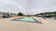 Located at 500 N Judge Ely Blvd in Abilene, Texas, this outdoor recreational area centers around a generously sized swimming pool with a classic rectangular design and metal handrail entry points. The pool deck is finished in a light-toned, textured surface that surrounds the water on all sides, providing ample space for relaxation and movement. Vibrant orange lounge chairs are arranged along the poolside, offering comfortable spots for sunbathing and leisure. Multiple green-painted pergola structures are positioned throughout the area, providing shaded seating with outdoor tables and chairs beneath them — ideal for gathering or simply escaping the Texas sun. A gated perimeter with wrought iron-style fencing encloses the space, offering both security and a defined boundary that separates the pool area from the surrounding property. Mature trees visible beyond the fence add a natural backdrop, while the low-rise building architecture of the surrounding complex is visible in multiple directions. The open-air layout and spacious deck make this an inviting outdoor amenity well-suited for community use, offering both active recreation in the pool and relaxed social spaces under the pergolas.