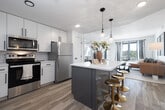 This thoughtfully designed 2-bedroom, 2-bathroom residence offers a cohesive modern aesthetic across its single floor. The open-concept kitchen and living area serves as the heart of the home, featuring white shaker cabinetry, subway tile backsplash, stainless steel appliances, and a gray kitchen island with a white quartz countertop and gold bar stools beneath elegant pendant lighting. Light wood-style flooring flows throughout, enhancing the bright, airy feel of the space. The living room is generously sized, bathed in natural light from large windows, and provides views of the outdoors. Both bedrooms offer carpeted floors, natural light, and a calm, restful atmosphere. The primary bedroom includes access to a walk-in closet with built-in shelving and hanging rods. Each bathroom is finished with gray vanities, white countertops, black hardware accents, and wood-look flooring, with one bathroom featuring a tub-shower combo. A dedicated closet provides additional storage. Throughout the residence, clean white walls, consistent finishes, and a neutral color palette create a polished, move-in-ready environment suited for comfortable everyday living.