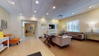 This single-floor facility is thoughtfully designed to support both relaxation and an active lifestyle across several distinct communal spaces. The main lounge serves as a welcoming entry point, featuring warm yellow-toned walls, large windows that admit natural light, and tile flooring that transitions to carpeted seating areas. Comfortable upholstered sofas and a dark wood coffee table are arranged around a wall-mounted television, with decorative wall art and fresh floral accents contributing to the inviting atmosphere. A corridor leads further into the building, connecting to additional amenities.

The expansive common room offers generous open space with recessed lighting set into a drop-panel ceiling. Dark wood high-top dining tables and chairs are arranged throughout, alongside upholstered seating clusters near elevator access, making it suitable for both casual gatherings and dining. The neutral carpet and tile flooring combination provides a clean, versatile aesthetic.

The fitness center is well-appointed with rubber flooring and includes cardio equipment such as treadmills and ellipticals, as well as a free-weight station with a dumbbell rack, exercise balls, and a water fountain. Wall-mounted televisions provide entertainment during workouts. Together, these spaces create a balanced, functional environment designed to serve a community of residents.