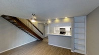 This two-story space features an open-concept main level that seamlessly connects the living area and kitchen. The living area is finished with wood-look laminate flooring and neutral gray walls, creating a clean, modern aesthetic. A textured ceiling with recessed lighting provides warm, even illumination throughout the space, complemented by a pendant light above the staircase landing.

The kitchen is accessible via a raised pass-through counter, offering both functionality and an open sightline to the living area. White cabinetry lines the kitchen walls, and black appliances, including a range, add a sharp contrast. Built-in open shelving on one wall provides additional storage and display space.

A wood-railed staircase with warm-toned treads rises along one wall, leading to the second floor. The staircase adds architectural interest to the main level while maximizing the use of vertical space. The overall design emphasizes practicality and a cohesive, neutral color palette, making the interior feel bright and well-organized throughout both levels.