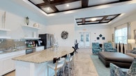 This single-floor social room is designed to foster community and comfort, blending functional amenities with sophisticated design elements. The space is anchored by a striking granite-topped island bar with seating, making it an ideal gathering spot for casual conversations or events. Adjacent to the island, a fully equipped kitchen features stainless steel appliances, including a French-door refrigerator, white cabinetry, and ample counter space, providing everything needed for entertaining.

The lounge area transitions seamlessly from the kitchen, offering plush seating that includes patterned accent chairs and a sofa, arranged to encourage relaxed socializing. Light wood-toned flooring runs throughout, lending warmth and continuity to the entire space. A workstation area near the entrance provides a functional nook for residents needing a quiet place to focus.

Overhead, elegant coffered ceilings with dark wood framing and recessed lighting create a refined architectural focal point that elevates the room's overall ambiance. French doors near the lounge area welcome natural light and provide a connection to the exterior. Decorative wall accents and carefully chosen furnishings in teal and neutral tones complete the polished, welcoming aesthetic of this thoughtfully appointed social space.
