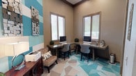 This thoughtfully designed business center offers a functional yet visually striking workspace for residents or guests. The room is anchored by an L-shaped, wraparound desk finished in a warm gray tone, providing ample workspace accommodating two desktop computer workstations side by side. Upholstered task chairs with a neutral finish complement the practical setup, while small potted greenery on the desktop adds a refreshing, organic touch.

Natural light fills the space through two tall windows dressed with horizontal blinds, offering both brightness and privacy control. The elevated ceiling height enhances the sense of openness within the compact room.

The décor is a standout feature, with a bold teal and white horizontal stripe accent wall paired with a geometric mosaic wall art piece, lending the space a contemporary and creative energy. The geometric-patterned floor tiles echo the color palette, blending teal, white, and taupe tones in an eye-catching design that ties the room together.

A comfortable upholstered armchair with decorative pillows occupies one corner, offering a relaxed seating option. A printer station is conveniently positioned nearby, ensuring all essential office amenities are within reach. Overall, the space balances professional utility with modern, curated design.