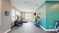 This dedicated fitness center is a comprehensively outfitted space designed to support a full range of workout routines. The layout is thoughtfully organized, with the main workout area featuring durable, dark rubberized flooring suited for high-activity use. A bold teal accent wall adds a vibrant visual element that energizes the space and contrasts beautifully against the neutral beige walls throughout. Recessed lighting in the white ceiling provides consistent illumination, while multiple windows fitted with blinds allow ample natural light to filter in and offer views of the surrounding neighborhood. The cardio zone is equipped with a treadmill and an elliptical stair climber, positioned near the windows to make workouts more engaging. A multi-functional cable trainer station anchors one section of the room, accompanied by wall-mounted resistance accessories. A vertical ball rack displays an assortment of medicine balls, stability balls, and resistance bands, keeping equipment neatly organized. A dumbbell rack adds further versatility for strength training. An adjacent hallway with hardwood flooring connects to additional rooms, suggesting changing or utility facilities nearby. An exterior door with an exit sign provides direct outdoor access, adding convenience and safety to the overall design.