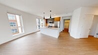 This contemporary 1,100-square-foot apartment in Redmond, WA features 2 bedrooms and 2 bathrooms across a single floor, delivering a clean, modern aesthetic throughout. The open-concept kitchen and living area serves as the heart of the home, anchored by a large quartz waterfall island with an undermount sink and sleek pendant lighting. The kitchen is outfitted with two-tone cabinetry in warm wood and matte gray finishes, a white subway tile backsplash, and a full suite of stainless steel appliances including a refrigerator, range, and microwave. Light wood-look flooring flows seamlessly throughout the unit. The living area is bathed in natural light from oversized windows offering views of the surrounding Redmond neighborhood. The primary bedroom measures 140 square feet and features a ceiling fan and large windows. The second bedroom is equally bright with a ceiling fan and a wide window framing outdoor views. The primary bathroom includes a glass-enclosed walk-in shower with large-format white tile and matte black fixtures, while the second bathroom features a soaking tub with a tiled surround and warm wood vanity. Additional spaces include a dedicated laundry area, closet, and hallway, rounding out this well-appointed modern residence.