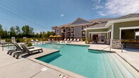 Located at 2811 Technology Dr in Lake Saint Louis, Missouri, this outdoor amenity area is designed to deliver a resort-like experience. The centerpiece is a generously sized swimming pool with crystal-clear water, complemented by a separate hot tub/spa for relaxation. A standout feature is the set of in-water sun loungers positioned directly in the shallow pool area, offering a unique way to cool off while soaking up the sun.

Surrounding the pool is an expansive concrete pool deck furnished with multiple chaise loungers and shaded seating areas with vibrant yellow umbrellas, providing ample space for sunbathing and socializing. The deck flows seamlessly toward a covered outdoor pavilion, which features stone accents, an outdoor fireplace, ceiling fans, and large glass doors — creating a comfortable indoor-outdoor lounge and dining area usable across seasons.

The two-story residential building rises behind the pool area, featuring craftsman-inspired architecture with board-and-batten siding, brick detailing, pitched rooflines, and private balconies overlooking the pool. The entire area is enclosed by a decorative iron fence, with lush greenery visible beyond. The combination of thoughtful amenities, architectural detail, and open sky creates a bright, welcoming outdoor retreat ideal for leisure and community gathering.