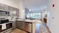 This well-appointed one-bedroom, one-bathroom residence at Quarry Hills in Quincy, MA offers a thoughtfully designed open-concept layout across a single floor. The kitchen serves as a central hub, featuring granite countertops, contemporary wood-tone cabinetry, stainless steel appliances including a dishwasher and refrigerator, and a peninsula with pendant lighting overhead — ideal for both meal preparation and casual dining. The kitchen flows seamlessly into the living room, which is highlighted by soaring ceilings, warm hardwood-style flooring, and a large picture window that invites abundant natural light and outdoor views. A dedicated closet provides convenient storage within the unit. The spacious primary bedroom offers plush carpeting, a window with exterior views, and elevated ceilings that contribute to an airy feel. The full bathroom is finished with grey subway tile surrounding a soaking tub and shower, a wood-tone vanity with a white countertop, and tile flooring, creating a clean and modern aesthetic. Throughout the residence, a cohesive color palette of soft blues, warm neutrals, and crisp whites establishes a calm and contemporary ambiance, making this an efficient and comfortable living space.