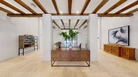 This expansive single-floor space spans a generous footprint, thoughtfully designed to blend rustic architectural character with contemporary, curated interiors. Exposed wood ceiling beams are a defining feature throughout, adding warmth and texture against crisp white walls and soaring ceiling heights that lend the space an airy, open feel.

The entrance greets visitors with large glass doors framed in dark metal, flooding the foyer with natural light and views of lush outdoor greenery. A striking chandelier anchors the entry, setting an elegant tone. Marble tile flooring transitions into warm hardwood in the interior zones, clearly delineating different areas of the space.

The living room is generously proportioned, furnished with plush white sectional seating arranged around a trio of wall-mounted screens on a bold charcoal accent wall, complemented by a coffered ceiling with recessed lighting. A separate, more intimate lounge area features a mirrored backdrop and a sculptural wood credenza adorned with potted greenery.

The kitchen and dining area exudes a relaxed coastal aesthetic, centered on a large island with patterned tile detailing and bar seating beneath cascading pendant lights. A built-in banquette with bold striped cushions and a woven pendant light creates a cozy dining nook with natural light from a nearby window, completing the inviting, well-appointed atmosphere.