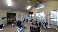 This fitness center is designed to provide a functional and inviting workout environment. The space is defined by dramatic vaulted ceilings that create a sense of openness and volume, complemented by a combination of skylights and large multi-pane windows — including a striking arched window — that flood the room with natural light and offer pleasant views of the surrounding greenery outside.

The facility is outfitted with a comprehensive selection of exercise equipment to accommodate a variety of fitness routines. Cardio machines, including a stationary bike and an elliptical trainer, are positioned throughout the space, while a dedicated strength training area features a multi-station cable machine, a weight bench with rack, and a full dumbbell rack with an assortment of free weights. A wall-mounted flat-screen television is conveniently positioned for use during workouts.

A wall of mirrors enhances the sense of space and supports proper form during exercise. The flooring incorporates protective rubber matting in key equipment areas. An adjoining room, visible through a doorway, appears to offer additional amenities. The overall ambiance is bright, airy, and well-maintained, making this fitness center a practical and welcoming resource for residents or users of the facility.