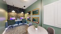 Located at 300 Gatehouse Ln in Odenton, Maryland, this business center offers a thoughtfully designed, single-floor workspace suited for both focused productivity and casual collaboration. The space is distinguished by its rich sage green walls, which create a calm and professional atmosphere throughout. A standout feature is the textured stone accent wall at the workstation area, adding an earthy, contemporary character to the room. Built-in desk stations are equipped with desktop computers and illuminated by pendant lights overhead, providing a well-lit environment for individual work. Patterned, neutral-toned carpeting runs throughout, adding warmth and visual interest underfoot. A round table with modern seating serves as a collaborative gathering spot, ideal for informal meetings or group discussions. Four wood-framed mirrors are artfully arranged on the green accent wall, enhancing the sense of space and light. Natural light enters through a window fitted with blinds, offering a connection to the outdoors. An adjacent room, visible through a doorway, features additional lounge-style seating and decorative lighting, extending the functional footprint of the space. Clean white trim and millwork details complete the polished, cohesive aesthetic of this well-appointed business center.