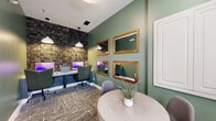 Located at 300 Gatehouse Ln in Odenton, Maryland, this business center offers a thoughtfully designed, single-floor workspace suited for both focused productivity and casual collaboration. The space is distinguished by its rich sage green walls, which create a calm and professional atmosphere throughout. A standout feature is the textured stone accent wall at the workstation area, adding an earthy, contemporary character to the room. Built-in desk stations are equipped with desktop computers and illuminated by pendant lights overhead, providing a well-lit environment for individual work. Patterned, neutral-toned carpeting runs throughout, adding warmth and visual interest underfoot. A round table with modern seating serves as a collaborative gathering spot, ideal for informal meetings or group discussions. Four wood-framed mirrors are artfully arranged on the green accent wall, enhancing the sense of space and light. Natural light enters through a window fitted with blinds, offering a connection to the outdoors. An adjacent room, visible through a doorway, features additional lounge-style seating and decorative lighting, extending the functional footprint of the space. Clean white trim and millwork details complete the polished, cohesive aesthetic of this well-appointed business center.