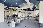Located on South University Avenue in Ann Arbor, Michigan, this single-floor fitness facility offers an expansive, well-appointed space designed to support a wide range of workout modalities. The main gym floor features a dark navy exposed ceiling with white structural ductwork, large ceiling fans, and distinctive yellow pendant lighting that adds a modern industrial character. The flooring throughout the equipment areas is durable rubber gym tile, providing both safety and functionality. The strength and conditioning zone is comprehensively equipped with free weights, a full dumbbell rack, weight benches, cable machines, and multi-use strength equipment, all arranged around wall-to-wall mirrors that enhance the sense of space. The cardio zone is lined with rows of treadmills and elliptical machines from Matrix, positioned in front of large windows that frame views of the surrounding Ann Arbor streetscape, flooding the area with natural light. A dedicated open studio room stands apart with its striking yellow geometric ceiling feature, light hardwood floors, floor-to-ceiling windows offering panoramic city views, decorative wall art, and integrated lighting — ideal for group fitness, yoga, or movement classes. Throughout, the facility maintains a clean, modern aesthetic with an energetic and motivating ambiance.