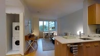 This well-designed single-floor residence offers a seamless open-plan layout that integrates the kitchen, dining, and living areas into one cohesive and functional space. The kitchen features a central island with a clean white countertop and warm wood cabinetry, providing both practical workspace and a natural visual anchor for the room. Light hardwood flooring runs throughout, lending warmth and continuity across the space.

The dining area is positioned directly in front of large floor-to-ceiling windows that flood the interior with natural light and offer views of the surrounding urban greenery and neighbouring buildings. A compact dining table paired with industrial-style metal chairs creates an understated yet stylish setting. Adjacent to the dining area, a dedicated workspace with a wooden fold-out desk and chair offers a practical nook for focused tasks.

The living area is comfortably arranged with a sofa and soft furnishings, complemented by a striking arc floor lamp that adds a sculptural element to the room. A area rug grounds the seating zone and adds texture to the space. The overall ambiance is calm and contemporary, with a neutral colour palette of whites and soft greys that enhances the sense of openness and light throughout.