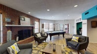 This single-floor community space is thoughtfully designed to encourage socializing and relaxation, blending functional amenities with a warm, eclectic aesthetic. The main gathering area is anchored by a full-size pool table positioned at its center, surrounded by generous seating arrangements that include upholstered sofas and accent chairs in neutral tones, accented with yellow and gray throw pillows. A bold, geometric-patterned area rug defines the lounge zone beneath a wooden coffee table.

A prominent brick fireplace with a raised hearth serves as a focal point along one wall, adding a rustic, grounded character to the space. Decorative signage and wall art contribute to the inviting atmosphere. Multiple windows allow natural light to flow throughout, and recessed ceiling lighting provides consistent illumination.

An adjacent nook features a vibrant teal accent wall, wall-mounted flat-screen television, and comfortable seating, offering a more intimate retreat within the larger space. A compact but well-appointed kitchenette is also present, outfitted with warm wood cabinetry, granite-style countertops, a mosaic tile backsplash, and stainless steel appliances including a range, microwave, refrigerator, and sink. Light wood-style flooring runs throughout, unifying the space with a clean, cohesive finish.