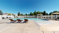 This outdoor pool facility in West Jordan, Utah offers a well-appointed recreational space designed for leisure and relaxation. The centerpiece is a generously sized swimming pool with clear, blue water, complemented by a shallow wading area that gradually slopes from the pool's edge — ideal for a range of swimmers. Adjacent to the main pool sits a hot tub, providing an additional amenity for relaxation. The expansive concrete sun deck surrounds the pool and provides ample space for lounging, furnished with multiple slatted chaise lounge chairs positioned to take advantage of Utah's sunny climate. White vinyl fencing encloses the entire area, ensuring privacy and safety, while mature trees lining the perimeter create a naturally shaded backdrop. Two white pergolas are positioned within the fenced area, offering shaded seating and a pleasant gathering spot. A dedicated pool house structure features stone veneer accenting at its base, complementing the neutral stucco exterior — a design detail echoed throughout the facility's architecture. The surrounding landscape, featuring lush green trees and open sky views, enhances the overall outdoor ambiance, making this pool facility a standout community amenity.