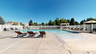 This outdoor pool facility in West Jordan, Utah offers a well-appointed recreational space designed for leisure and relaxation. The centerpiece is a generously sized swimming pool with clear, blue water, complemented by a shallow wading area that gradually slopes from the pool's edge — ideal for a range of swimmers. Adjacent to the main pool sits a hot tub, providing an additional amenity for relaxation. The expansive concrete sun deck surrounds the pool and provides ample space for lounging, furnished with multiple slatted chaise lounge chairs positioned to take advantage of Utah's sunny climate. White vinyl fencing encloses the entire area, ensuring privacy and safety, while mature trees lining the perimeter create a naturally shaded backdrop. Two white pergolas are positioned within the fenced area, offering shaded seating and a pleasant gathering spot. A dedicated pool house structure features stone veneer accenting at its base, complementing the neutral stucco exterior — a design detail echoed throughout the facility's architecture. The surrounding landscape, featuring lush green trees and open sky views, enhances the overall outdoor ambiance, making this pool facility a standout community amenity.