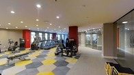 Located at 4451 Telfair Blvd in Camp Springs, Maryland, this well-appointed fitness center offers a comprehensive workout environment across a single level. The space is designed with functionality and modern aesthetics in mind, featuring recessed lighting throughout that keeps the area bright and energizing.

The main workout floor showcases a row of cardio machines, including treadmills and ellipticals, positioned along natural-light-admitting windows that line one wall. Complementing these are a variety of strength and resistance training machines arranged across the open floor plan, providing ample space for a full-body workout routine.

The flooring is a distinctive geometric patterned carpet in shades of yellow, grey, and neutral tones, adding a bold, contemporary visual element to the space. A striking red accent column serves as a design focal point, anchoring the room and adding a vibrant contrast to the neutral ceiling and walls.

A glass-walled studio room is accessible from the main floor, offering a semi-private area ideal for group fitness classes, stretching, or personal training sessions. The transparency of the glass walls maintains an open, connected feel throughout the facility. Overall, the space is clean, organized, and thoughtfully designed to support a wide range of fitness activities.