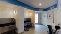 This single-floor space is purpose-built for pet washing and grooming, combining a functional design with a clean, welcoming aesthetic. The interior is defined by white subway tile walls that extend throughout, lending a sanitary, polished look while offering easy maintenance. A bold teal-blue upper wall color adds a playful contrast to the bright white tile below, creating a lively and approachable atmosphere.

The space features built-in pet bathing stations recessed within tiled alcoves, each equipped with a deep stainless steel tub, handheld fixtures, and a raised drainage grate on the floor. Industrial-style bar stools are positioned at each station for user convenience. Wall-mounted light fixtures provide warm, focused illumination within the bathing bays.

Recessed ceiling lighting brightens the overall space, which is finished with large-format light-toned tile flooring throughout. A glass entry door allows natural light to filter into the interior, contributing to an open feel. Decorative artwork featuring pet-themed illustrations adds a friendly, lighthearted touch to the walls. The overall design balances utility and charm, making it a practical yet inviting environment for pet care services.