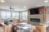 Located at 6604 Odana Rd in Madison, WI, this generously sized single-floor amenity space is thoughtfully designed to serve as a social and gathering hub. The open-concept interior seamlessly connects a fully equipped community kitchen and a comfortable lounge area, all unified by warm hardwood-style flooring and recessed lighting throughout.

The kitchen is a standout feature, anchored by a large granite-topped island with bar seating for multiple guests. Stainless steel appliances — including a refrigerator, range, microwave, and dishwasher — line the wall beneath a striking herringbone-patterned tile backsplash. A bold teal ceiling accent and industrial pendant lighting add a modern design flair overhead.

The adjacent lounge area is inviting and well-appointed, centered around a linear gas fireplace set against a warm wood-paneled accent wall with a wall-mounted television above. Generous seating arrangements, ceiling fans, and abundant natural light from multiple windows create a relaxed, comfortable atmosphere.

The space extends outdoors to a large elevated terrace offering panoramic views of the Madison skyline and surrounding greenery. The terrace includes a covered grilling station with built-in BBQs, a fire pit lounge area with cushioned seating, and additional dining arrangements — ideal for outdoor gatherings in any season.