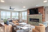 Located at 6604 Odana Rd in Madison, WI, this generously sized single-floor amenity space is thoughtfully designed to serve as a social and gathering hub. The open-concept interior seamlessly connects a fully equipped community kitchen and a comfortable lounge area, all unified by warm hardwood-style flooring and recessed lighting throughout.

The kitchen is a standout feature, anchored by a large granite-topped island with bar seating for multiple guests. Stainless steel appliances — including a refrigerator, range, microwave, and dishwasher — line the wall beneath a striking herringbone-patterned tile backsplash. A bold teal ceiling accent and industrial pendant lighting add a modern design flair overhead.

The adjacent lounge area is inviting and well-appointed, centered around a linear gas fireplace set against a warm wood-paneled accent wall with a wall-mounted television above. Generous seating arrangements, ceiling fans, and abundant natural light from multiple windows create a relaxed, comfortable atmosphere.

The space extends outdoors to a large elevated terrace offering panoramic views of the Madison skyline and surrounding greenery. The terrace includes a covered grilling station with built-in BBQs, a fire pit lounge area with cushioned seating, and additional dining arrangements — ideal for outdoor gatherings in any season.